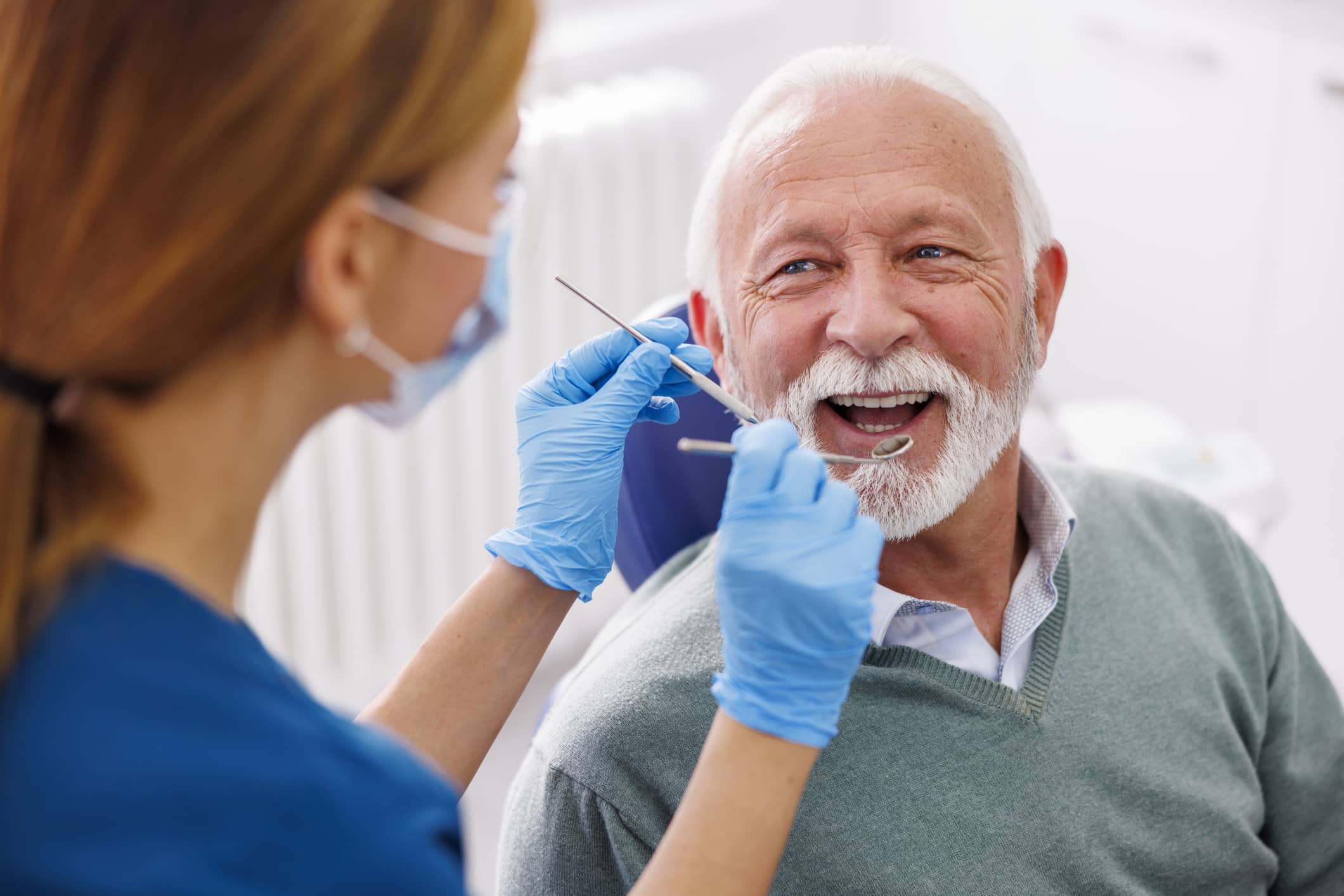Elderly man with healthy teeth smiling at dental technician as they approach his mouth with dental tools.